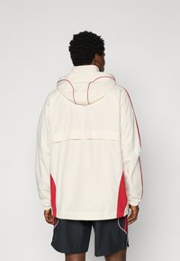 White hooded jacket with red side accents and a drawstring hem. Smooth texture, loose fit, and visible stitching details.