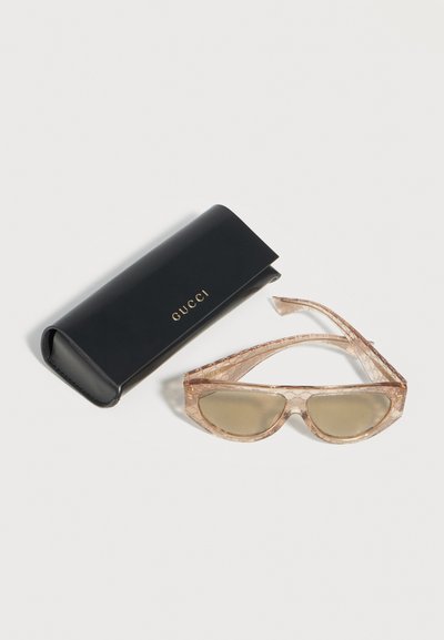 Διαφανή μπεζ γυαλιά ηλίου Gucci με διακριτικό μοτίβο λογότυπου δίπλα σε μαύρη σκληρή θήκη Gucci με χρυσά γράμματα σε ανοιχτό φόντο.