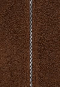 Brun fleecejakke med en tekstureret overflade. Har en sølvfarvet lynlås, der løber centralt og står i kontrast til den bløde stof.