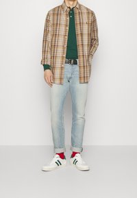 Camicia a quadri marrone e beige sopra una maglietta verde a maniche lunghe, abbinata a jeans blu chiaro e sneakers bianche con strisce verdi.