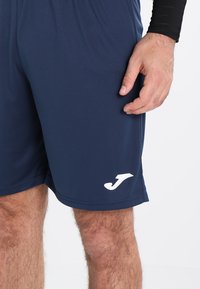 Marineblå træningsshorts lavet af letvægtsstof, med et hvidt logo og en elastisk talje for komfort. Glat tekstur.