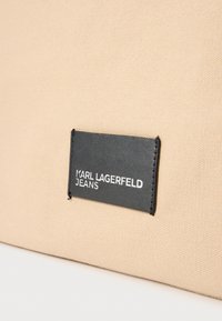 Smėlio spalvos drobinis maišas su juodu stačiakampiu lipduku, ant kurio reljefiškai išgraviruotas tekstas „KARL LAGERFELD JEANS“. Paprastas dizainas ir matinė tekstūra.