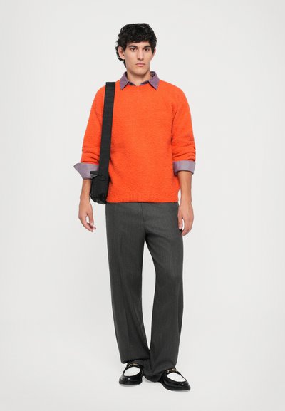 Orange tekstureret sweater, grå stribede bukser, sort crossover-taske og sorte sko med hvide accenter. Model står foran en hvid baggrund.