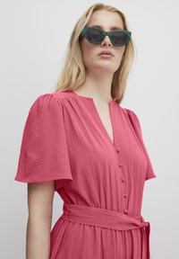 Abito rosa, con texture, maniche a palloncino corte, scollatura a V, bottoni frontali e una cintura abbinata legata in vita. Abbinato a occhiali da sole verdi.
