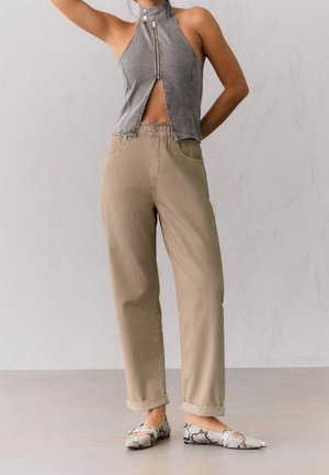 Femme portant un haut sans manches gris à fermeture éclair, un pantalon beige taille haute à jambes larges et des chaussures plates à motif peau de serpent, debout avec le bras levé.