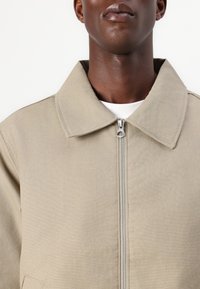 Beige zip-up jacket made of canvas, with a pointed collar and tonal stitching. Features a silver zipper and a white undershirt visible underneath.