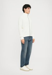 Muškarac stoji, odjeven u bijeli duksericu na zatvaranje, plave traperice i bijele tenisice, nasuprot jednostavne svijetle pozadine.