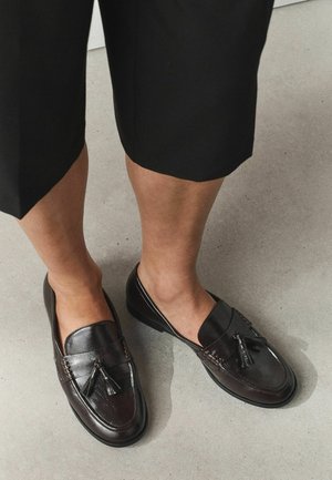 Beine, die schwarze, gekürzte Hosen und dunkelbraune Leder-Loafer mit Quasten tragen, stehen auf einem hellgrauen Boden.