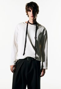 Weißes langärmliges Hemd mit lockerem Sitz, das einen schwarzen, strukturierten Riemen um den Hals aufweist, kombiniert mit schwarzen Hosen.