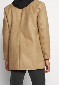 Manteau en mélange de laine couleur camel avec une coupe droite, des épaules structurées et une capuche noire. Présente des lignes épurées et un minimum de détails.