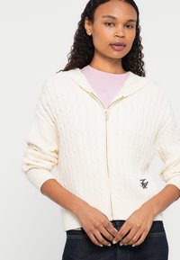 Kremowy, dziany hoodie z zamkiem błyskawicznym, z wzorem w warkocze, prążkowanymi mankietami i dołem. Posiada złoty zamek i małe haftowane logo po lewej stronie.