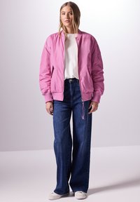 Różowa kurtka bombowca z ściągaczami, połączona z ciemnoniebieskimi dżinsami z wysokim stanem i szerokimi nogawkami oraz białymi trampkami. Prosta biała koszulka pod spodem.
