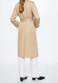 Trench-coat camel avec une ceinture à la taille, en tissu lisse. Présente un design classique, des manches longues, et descend jusqu'au milieu du mollet au-dessus d'un pantalon blanc.