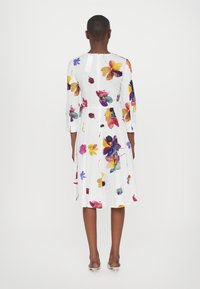 Valkoinen mekko, jossa on eläväinen kukkakuosi purppurassa, keltaisessa ja tealissa. Pitkät hihat, istuva vyötärö ja liehuva hame, jossa on laskoksia.