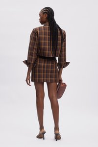 Set di giacca cropped a quadri marroni e mini gonna con dettagli di maniche arricciate. La modella tiene in mano una clutch terracotta e indossa sandali con tacco trasparente.