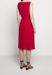 Červené skládací šaty bez rukávů s opaskem v pase a zipem na zadní straně; doplněné černými sandály na pásku pro kompletní vzhled.