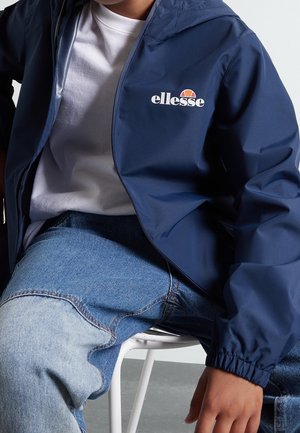 Granatowa wodoodporna kurtka z zamkiem błyskawicznym z przodu, elastycznymi mankietami oraz białym logo „ellesse”; noszona na białej koszulce i niebieskich dżinsach.