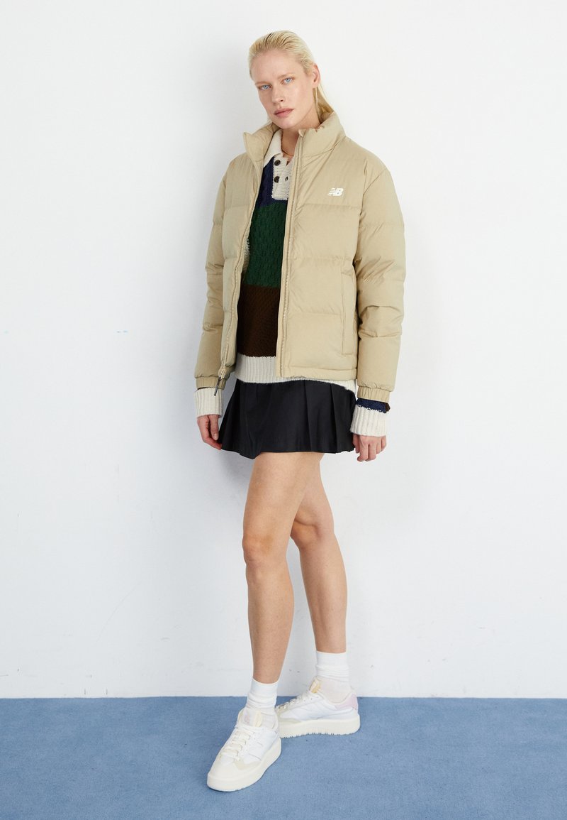 Veste en duvet beige avec fermeture éclair, poignets côtelés et logo. Superposée avec un pull en tricot vert et marron et une jupe plissée noire. Baskets blanches.