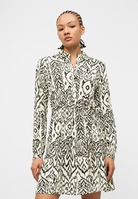 Γυναίκα με πλεξούδες που φοράει μακρυμάνικο φόρεμα με κουμπιά σε ασπρόμαυρο μοτίβο και ζώνη στη μέση.