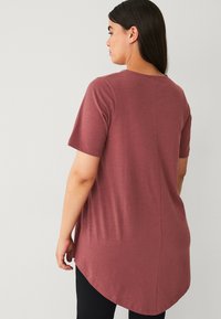 Bordeaux kortere tuniek met een afgeronde zoom en achternaad, gemaakt van zachte stof. Gemaakt over zwarte leggings, met een ontspannen pasvorm.