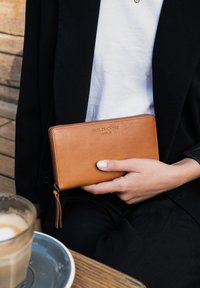 Tan leather wallet with a zipper, embossed logo, and a tassel. Held in a hand against a black jacket and a wooden background.
