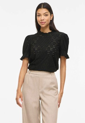 Mujer con una blusa negra de encaje de manga corta con hombros abullonados y pantalones beige de cintura alta, de pie frente a un fondo liso.