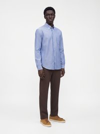 Homme debout portant une chemise bleue claire à boutons, un pantalon marron foncé et des baskets beiges sur un fond blanc uni.