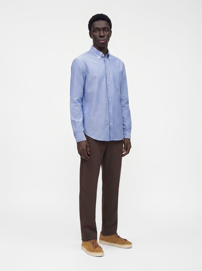 Homme debout portant une chemise bleue claire à boutons, un pantalon marron foncé et des baskets beiges sur un fond blanc uni.