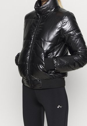 Schwarze glänzende Steppjacke mit Frontreißverschluss und elastischen Bündchen, getragen mit schwarzen Leggings mit kleinem weißem Logo am Oberschenkel.