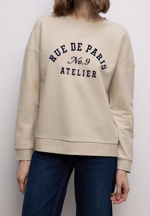 Beiger Pullover mit dem Text "RUE DE PARIS No.9 ATELIER", getragen mit dunkelblauer Jeans von einer Person, die von Hals bis Hüfte zu sehen ist.