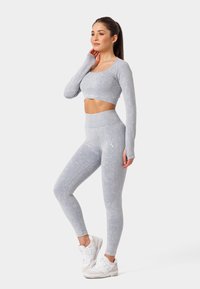 Gaiši pelēks piemērots garās piedurknes crop top un atbilstošas augstās jostas līnijas legingi, izgatavoti no mīksta, teksturēta auduma, kopā ar baltiem sporta apaviem.