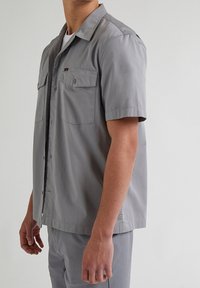 Maglietta grigia a maniche corte realizzata in tessuto liscio, con due tasche sul petto, chiusura con bottoni e un design del colletto ordinato.