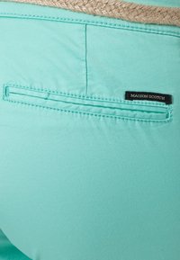 Pantalon vert menthe avec une poche arrière plate, présentant un accent tissé beigne au niveau de la taille et une étiquette noire portant l'inscription "MAISON SCOTCH."