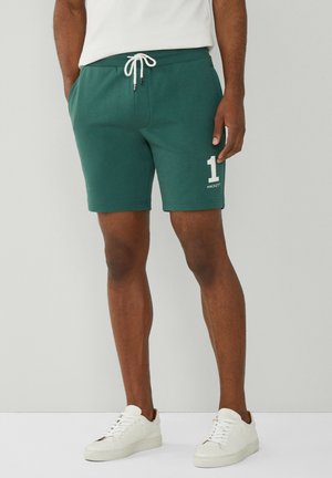 Man met donkergroene shorts met trekkoord, wit nummer "1" en tekst "Hackett" op het linkerbeen, gecombineerd met witte sneakers.