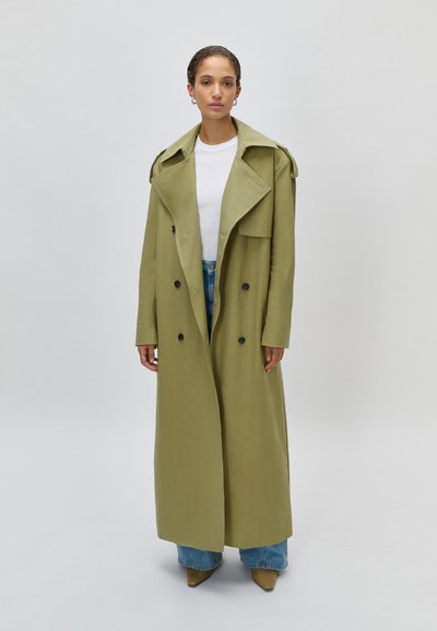 Γυναίκα που στέκεται, φοράει ένα μακρύ διπλόμπουτο παλτό σε ελιά πράσινο χρώμα, λευκό πουκάμισο, μπλε τζιν και καφέ μυτερές γόβες, μπροστά από ένα απλό φόντο.
