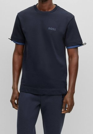 Homme portant une chemise bleu marine à manches courtes avec le logo "BOSS" sur la poitrine et un pantalon bleu marine assorti, sur un fond gris uni.