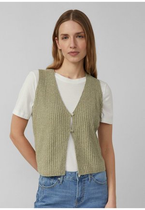 Kobieta o prostych, jasnobrązowych włosach, ubrana w oliwkową zapinaną na guziki, prążkowaną kamizelkę na białej koszulce z krótkim rękawem i niebieskich dżinsach, stojąca na szarym tle.