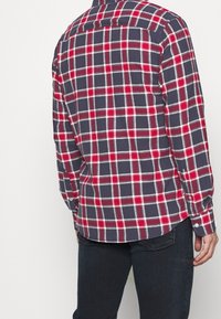 Camicia di flanella a quadri con un motivo plaid nei colori blu navy, rosso e bianco, maniche lunghe, polsini con bottoni e orlo curvo. Materiale in cotone.