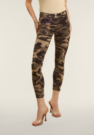 Nainen, joka käyttää lyhennettyjä maastokuvioituja housuja ja läpinäkyviä korkokenkiä, seisoo yksiväristä taustaa vasten.