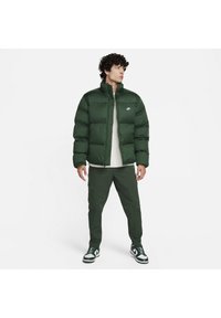 Kurtka puchowa w ciemnozielonym kolorze, z wysokim kołnierzem i zapięciem na zamek. Połączona z pasującymi zielonymi spodniami. Posiada białe logo Nike na klatce piersiowej.