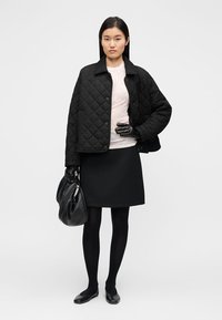 Μαύρο καπιτονέ μπουφάν με κουμπιά τύπου σναπ, γκρίζο τοπ, μαύρη φούστα, μαύρες καλσόν και γάντια, κρατώντας μια μαλακή μαύρη τσάντα.