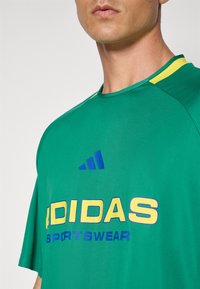 Zöld sportpóló sima anyagból, sárga és kék "ADIDAS SPORTSWEAR" logóval, kék díszcsíkokkal a vállakon.