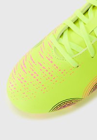 Lys gul fotballsko med teksturert overflate, med et gradientmønster i rosa og grønne lisser. Designet for ytelse på gressunderlag.