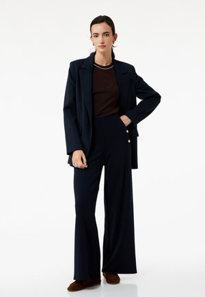 Mørkeblå blazer over en brun top, parret med vidde-bukser i mørkeblå, der har guldknapper i siden. Glat stof med tætsiddende pasform.