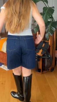 Personne aux longs cheveux roux portant un débardeur gris, un short en jean bleu et des bottes noires montantes aux genoux, debout à l'intérieur près d'une plante et d'un équipement d'exercice.
