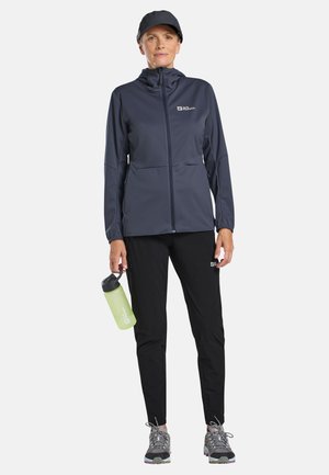 Vrouw in een marineblauw jack en zwarte broek houdt een groene waterfles vast, draagt grijze wandelschoenen en een zwarte pet, en staat tegen een witte achtergrond.