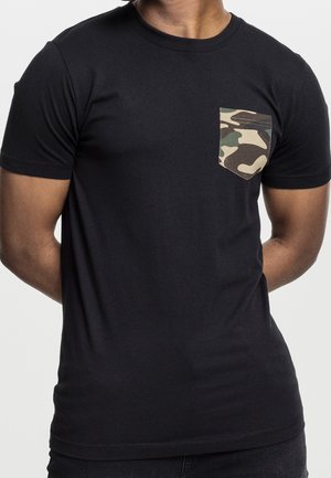 Mężczyzna ubrany w czarny krótki t-shirt z kieszenią na piersi w zielono-brązowy wzór kamuflażu.