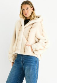 Young woman wearing a cream fur hooded jacket with hand in pocket and blue jeans against a white background.