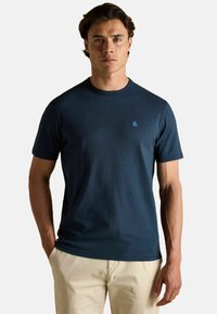 Jauns vīrietis valkā tumši zilu īsām piedurknēm T-kreklu ar mazu krūšu logo, apvienojot to ar bēšiem biksēm, stāvot uz baltā fona.