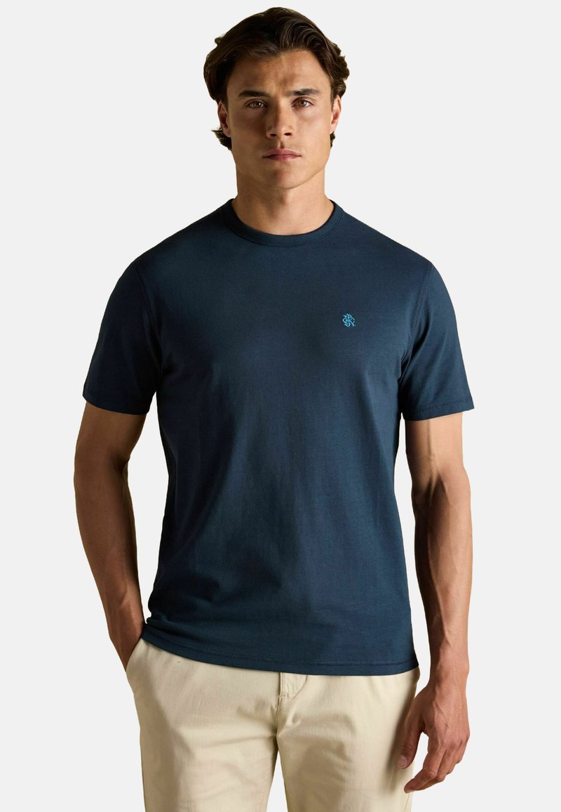 Jauns vīrietis valkā tumši zilu īsām piedurknēm T-kreklu ar mazu krūšu logo, apvienojot to ar bēšiem biksēm, stāvot uz baltā fona.